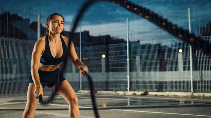 Beautiful Energetic Fitness Girl Doing Exercises with Battle Ropes. She is Doing a Workout in a Fenced Outdoor Basketball Court. Evening After Rain in a Residential Neighborhood Area.