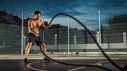 Fototapeta na wymiar Strong Muscular Fit Shirtless Young Man is Doing Exercises with Battle Ropes. He is Doing a Workout in a Fenced Outdoor Basketball Court. Evening After Rain in a Residential Neighborhood Area.