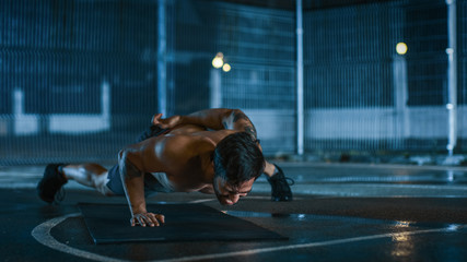 Strong Muscular Fit Shirtless Young Man is Doing One-Hand Push Up Exercises. He is Doing a Workout in a Fenced Outdoor Basketball Court. Night After Rain in a Residential Neighborhood Area.