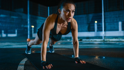Fototapeta premium Beautiful Sporty Fitness Girl Doing Push Up Exercises. She is Doing a Workout in a Fenced Outdoor Basketball Court. Night Footage After Rain in a Residential Neighborhood Area.