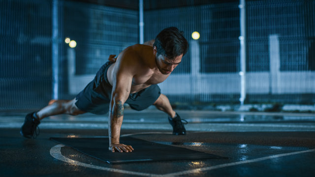 Strong Muscular Fit Shirtless Young Man is Doing One-Hand Push Up Exercises. He is Doing a Workout in a Fenced Outdoor Basketball Court. Night After Rain in a Residential Neighborhood Area. - Powered by Adobe