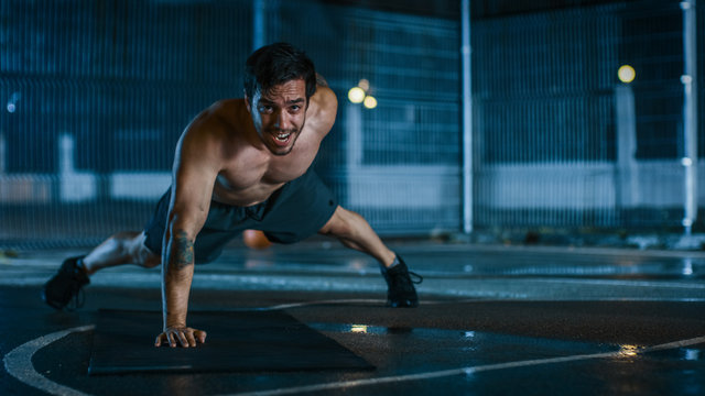 Strong Muscular Fit Shirtless Young Man is Doing One-Hand Push Up Exercises. He is Doing a Workout in a Fenced Outdoor Basketball Court. Night After Rain in a Residential Neighborhood Area.