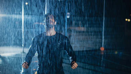 Strong Muscular Fit Young Man in Dark Athletic Clothes is Celebrating His Athletic Accomplishments. He is Cheering at Night in Heavy Rain with One Light Behind Him.