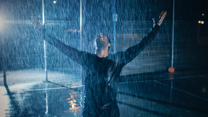 Strong Muscular Fit Young Man in Dark Athletic Clothes is Celebrating His Athletic Accomplishments. He is Cheering at Night in Heavy Rain with One Light Behind Him.