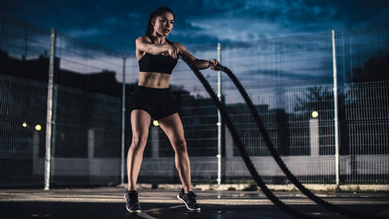 Fototapeta na wymiar Beautiful Energetic Fitness Girl Doing Exercises with Battle Ropes. She is Doing a Workout in a Fenced Outdoor Basketball Court. Evening After Rain in a Residential Neighborhood Area.