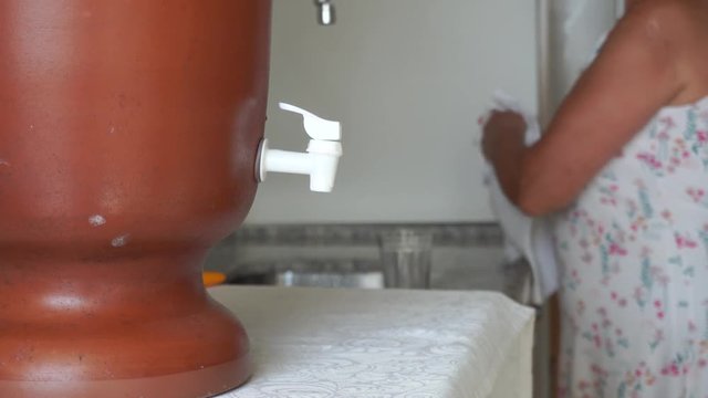 Old Woman at home is using a traditional clay water purifier to fill a glass of water em drinking it.