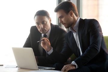 Serious focused businessmen in suits work together on online project management look at laptop discuss financial market, male partner makes business offer consult client talk give presentation advice