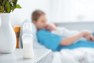close-up view of baby bottle with milk, vase and mother with newborn baby lying on hospital bed behind