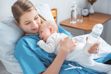 high angle view of smiling young mother holding baby bottle with milk and lying in bed with adorable baby