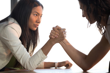 Two confident diverse businesswomen compete arm wrestling look in eyes feel jealous envious about...