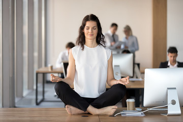 Calm young business woman doing yoga exercise at workplace sitting at work desk in lotus position, peaceful female employee office worker meditating relaxing taking break for stress relief concept