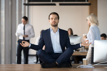 Mindful calm businessman in suit meditating at office sitting in lotus position on work desk,...