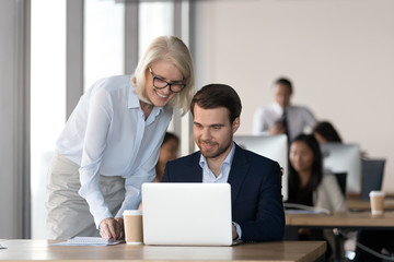 Friendly middle aged female mentor executive training employee in office helping with computer work, smiling old corporate manager supervise motivating teaching new worker colleague looking at laptop