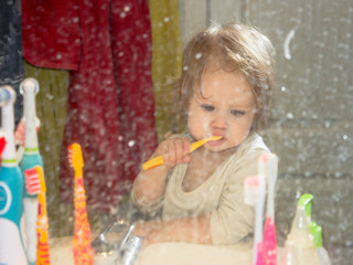 View through a dirty toothpaste-spattered mirror on a little baby girl. The child brushes his teeth, removes plaque from his first milk teeth in the bathroom, where everything is in disarray.
