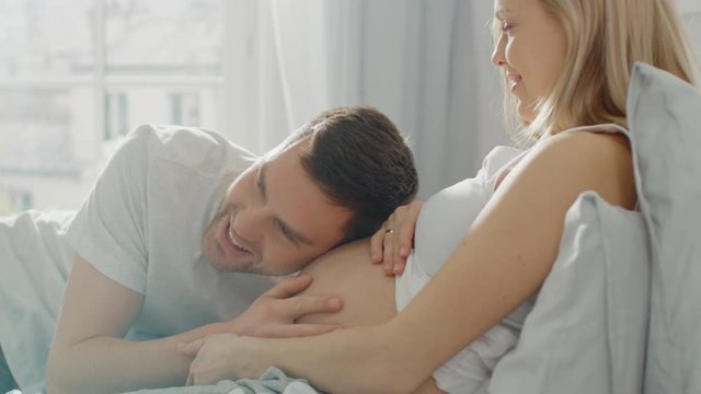 Happy Young Couple Cuddling Together in the Bed, Young Woman is Pregnant, Loving Partner Leans and Listens to a Baby Moving and it's Heartbeat in the Belly. Tender and Loving Moments of Family Life.