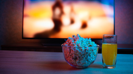 Popcorn in a glass plate with a drink on the background of the TV with the film on. Color bright...