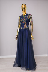 Fototapeta na wymiar Long, blue designer, evening, women's dress handmade on gold, glossy mannequin. With white and gold ornament in the form of a cherry tree with fruits from beads. Оn gray background.