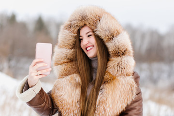 A beautiful girl in fur holds a phone in her hands and makes a selfie, a beautiful smile with white...
