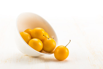 Handful of yellow cherry plums in ceramic bowl on white wooden surface
