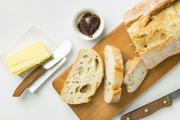 Breakfast table setting. Freshly baked artisan sliced bread on wood cutting board stick of organic butter homemade strawberry marmalade knife on white kitchen table. Top view. Morning energy
