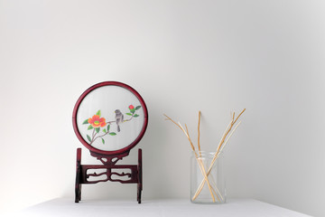 Vintage double sided glass Chinese silk embroidery mirror and glass vase with wicker rattan reed on white table top on white background in natural light with copy space Minimal Asian interior styling.