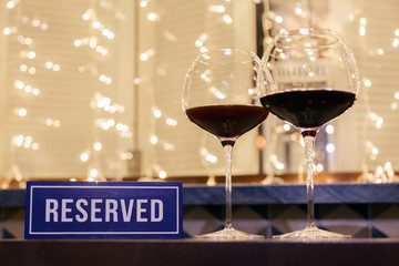 Closeup two crystal glasses red wine, reserved blue nameplate on wooden table, window, garlands, bokeh. Concept cozy evening in cafe, date, evening, Valentine Day, anniversary