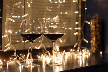 Closeup of two glasses of red wine in transparent crystal glasses on background of window and garlands. Concept of cozy evening in restaurant, date, christmas dinner, Valentine's Day