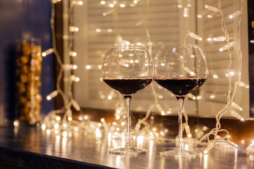 Closeup of two glasses of red wine in transparent crystal glasses on background of window, garlands. Concept of cozy evening in restaurant, date, christmas dinner, Valentine's Day