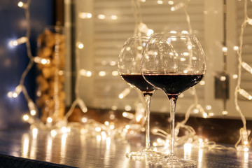 Closeup of two glasses of red wine in transparent crystal glasses on background of window, garlands. Concept of cozy evening in restaurant, date, christmas dinner, Valentine's Day