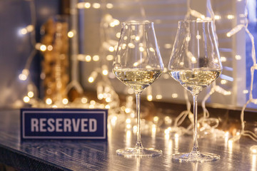 Closeup strawberry, two glasses white wine, reserved nameplate on wooden table, window, garlands, lights, bokeh. Concept date evening in restaraunt, 8 march, Valentine Day, christmas