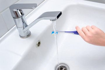 Close up image of a hand holding a blue toothbrush. water being poured on toothpaste on top in bathroom sink.