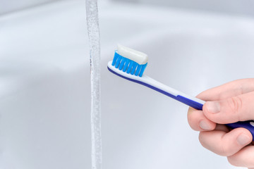 Close up image of a hand holding a blue toothbrush. water being poured on toothpaste on top in bathroom sink.