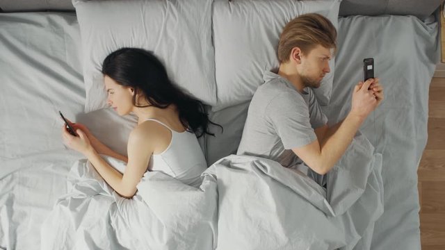 Alienated Millennial Young Couple in the Bed, Young People Turn Away From Each other Using Smartphones, Browsing Through Social Networks and Not Talking to Each Other. Top Down Camera Shot.