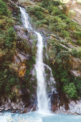 Fototapeta na wymiar A waterfall spotted in Tal, Annapurna Circuit Trek, Nepal. Few hundred meters of free fall, waterfall surrounded by tall mountains slopes, covered with green bushes and trees. Smooth capture