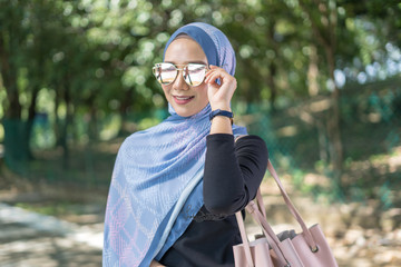 Portrait of attractive young Asian woman with tudung or headscarf and handbag posing at green park. Selective focus.