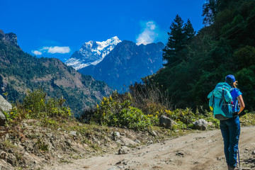 Obraz premium Trekking girl with a big blue backpack admires Manaslu, Annapurna Circuit Trek, Nepal. Forest to the right. To the left another Mountain. Manaslu covered with snow. Flowers next to the trekking trail.