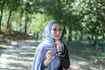 Portrait of attractive young Asian woman with tudung or headscarf and handbag posing at green park....