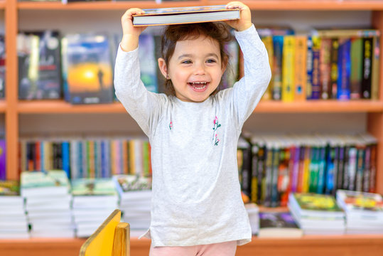 Cute Young Toddler Standing And Holding Book In Head. Little Happy Laughing Girl Indoors In Front Of Colorful Books. Child In A Library, Shop,Bookstore.