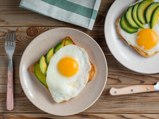 Toasts bread with fried eggs, avocado and cucumber slices on plates and wooden table with fork and knife. Top view