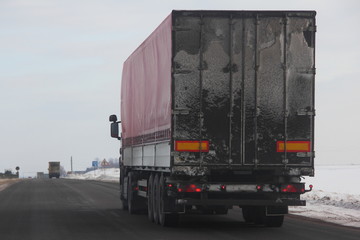 Logistics, international freight by road -  white european truck with semi trailer drive next to the two-lane asphalted suburban road in the winter day, rear view