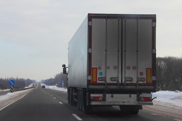 Logistics, international freight by road -  white european truck with semi trailer drive next to the two-lane asphalted suburban road in the winter day, back view