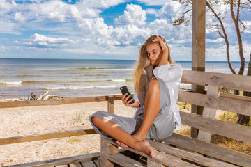 Fototapeta na wymiar Young girl with long fair hair sitting on a wooden pier on sea coast uses smartphone