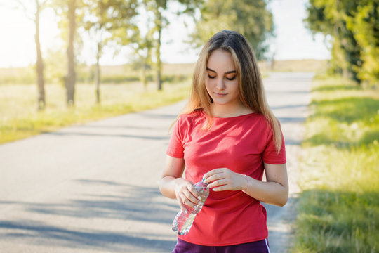 Young tenage girl in red t-shirt and violet shorts opening bottle of water against an evening sun. Beautiful sportsmen girl exhausted after evening running opens bottle of water