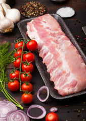 Plastic tray with raw pork ribs on chopping board and vintage meat fork and knife on wooden background. Fresh tomatoes and red onion with garlic, salt and pepper.