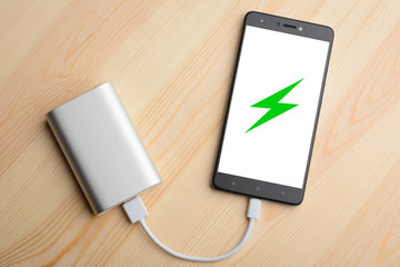 Modern smartphone lays on light wooden table while charging with power bank with quick charge. Illustration of green lightning is on the mobile phone screen