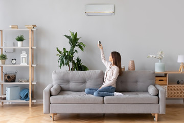 Young happy woman switching on air conditioner sitting on couch at convenient cozy home, lady relaxing on sofa in living room holding remote climate control to cooler system set comfort temperature