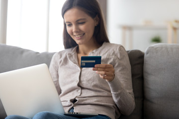 Happy young woman holding credit card using laptop for computer digital payment buying online, female customer making internet order purchase doing ecommerce shopping paying sitting on couch at home