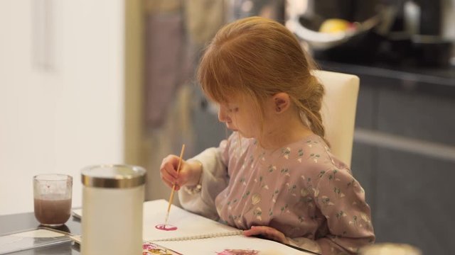 A beautiful little redhead girl draws pink circle in the notebook on kitchen table using a brush and water colours. The background is blurred.