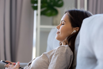 Serene calm young woman wearing earphones relaxing on sofa listening music on smartphone player...