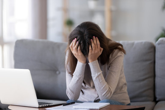 Upset millennial woman in panic holding head in hands frustrated shocked sitting in front on laptop on couch at home, tired bored student feel exhausted fatigued depressed having problem or headache